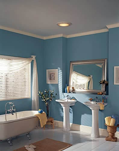 Round White Bath Exhaust Fan With Light And Glass Lens, 100 CFM installed in blue bathroom ceiling near mirror and sink area.