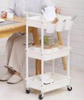 Three Tier Utility Rolling Cart With Lockable Caster Wheels And Three Hanging Baskets, shown in a dining setting, white finish.