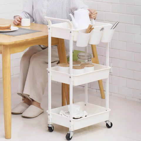 Three Tier Utility Rolling Cart With Lockable Caster Wheels And Three Hanging Baskets, shown in a dining setting, white finish.