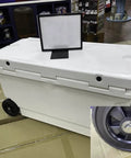 Large white rolling cooler with closed lid and visible wheel on store floor, displayed with a blank sign.