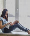 Woman sitting by window with baby, showcasing 48 X 102 Inch Fiberglass Window Screen Mesh for Windows and Doors Replacement Screen Door Mesh in Black Durable UV Protected Outdoor Screen.