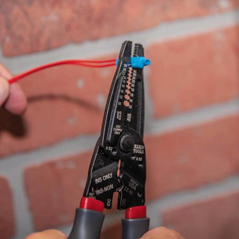 Kurve Wire Stripper Crimper Cutter Multi Tool for 10-26 AWG, cutting red wire against a brick wall, close-up of tool in use