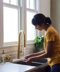 Woman washing hands at a kitchen sink with gold faucet and marble countertop, potted plant in background