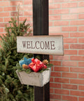Welcome sign with a rustic planter and red gift on a post against a brick wall background with greenery.