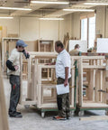 Workers in a carpentry workshop discussing wooden frames on dollies with heavy duty caster wheels, visible tools and machinery.
