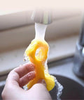 Hand washing a yellow silicone sponge under running water in a kitchen sink.