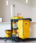 Heavy Duty 24 Gallon Front Zippered Vinyl Replacement Bag For Industrial Janitorial Carts Yellow Waste Linen And Trash Collection Durable Front Loading Bag For Commercial Cleaning Compatible With Rubbermaid Commercial Carts attached to a black cart alongside mops and other cleaning supplies in an office setting.